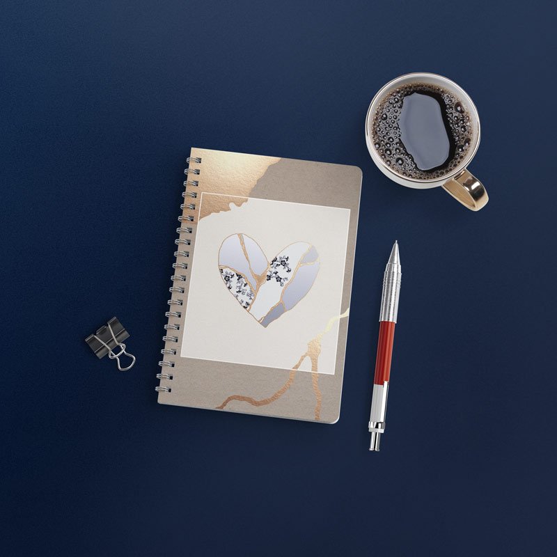 Birds eye view of a spiral notebook with illustrated heart on the cover, surrounded by coffee cup, pen, and paperclip on dark blue background..