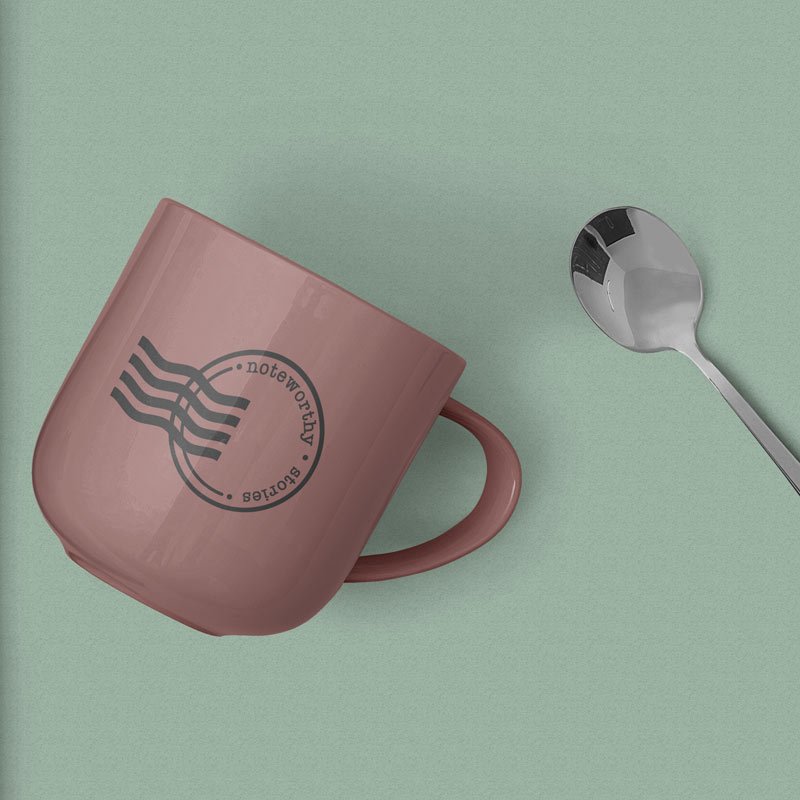 Noteworthy Stories basic logo printed on a rose pink mug with silver spoon beside it on a sage green background.